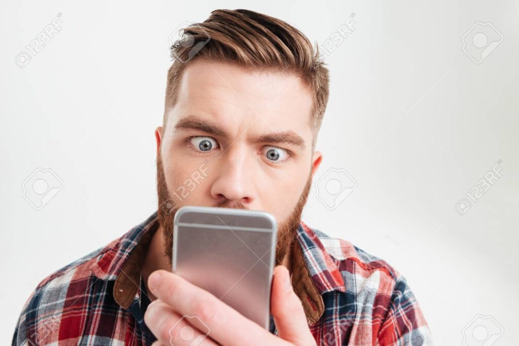 Close up portrait of a surprised casual man looking at mobile phone with big eyes over white background