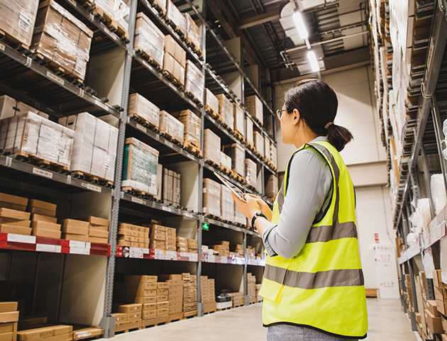 A female warehouse worker wearing a bright yellow high-visibility safety vest stands in an industrial warehouse aisle, holding a handheld scanner or mobile device. She has dark hair pulled back and is viewed from behind as she appears to be scanning or checking inventory. The warehouse features tall metal shelving units filled with cardboard boxes, pallets, and various stored goods organized on multiple levels. Fluorescent lighting illuminates the space from above, and the concrete floor and industrial ceiling structure are visible. The image represents modern warehouse operations, inventory management, and workplace safety protocols.