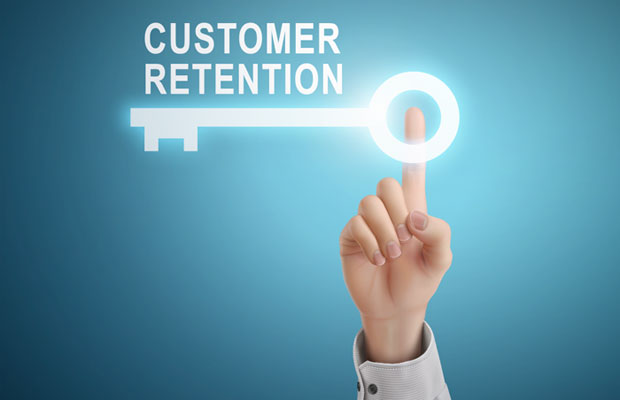 A human hand in a light-colored dress shirt sleeve extends upward against a blue gradient background, with the index finger touching a glowing white key icon. The text 'CUSTOMER RETENTION' appears in large white letters to the left of the key. The key has a circular head with a small human figure silhouette inside it. The image conceptually represents customer retention as the key to business success, with the interactive gesture suggesting unlocking or accessing customer loyalty strategies.
