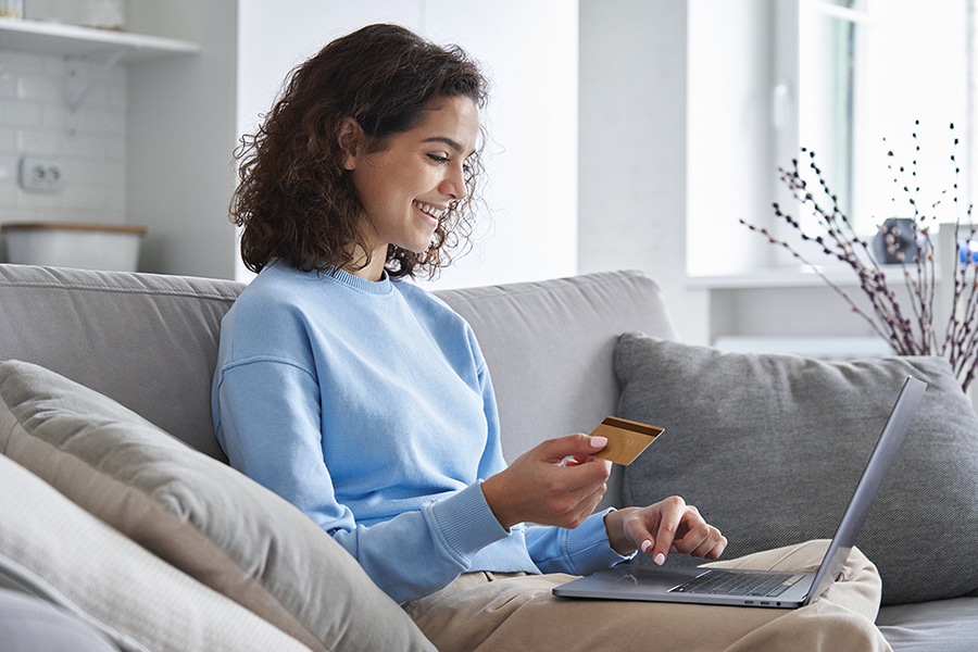 A young woman with curly brown hair sits comfortably on a light gray sofa in a bright, modern living room. She's wearing a light blue sweater and smiling as she holds a gold credit card in one hand while typing on a laptop computer with the other. The setting suggests she's making an online purchase from home. Behind her, white kitchen cabinets and decorative branches in a vase are visible, creating a cozy domestic atmosphere. The image represents online shopping, e-commerce, or digital banking from the comfort of home.
